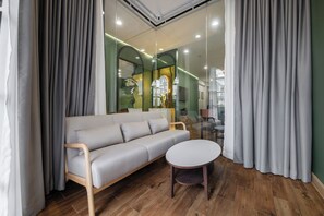 Una sala moderna con un sofá y una mesa redonda, suelo de madera y un separador de vidrio con vista a una tienda.