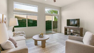Una sala de estar con sofá, una mesa de centro, televisor sobre un mueble bajo y ventanas amplias.