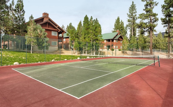 Un court de tennis avec un filet, entouré d’une clôture, et un bâtiment avec un toit végétalisé en arrière-plan.