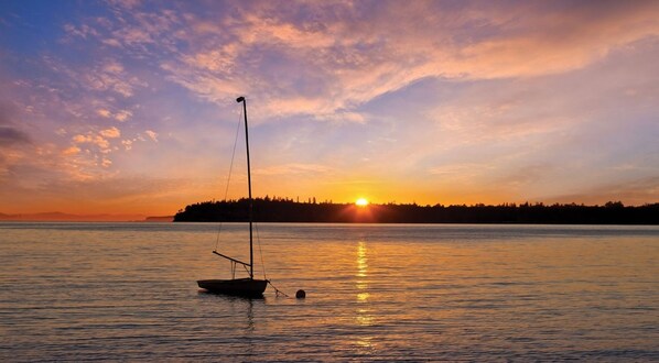 A calm body of water with a sailboat anchored near the shore, a sunset in the background, and a forested landmass across the water.
