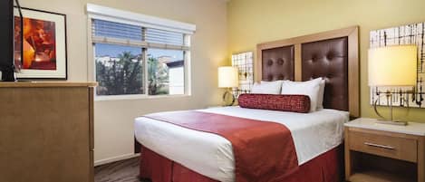 A hotel room with a large bed, a wooden nightstand, a desk, and a window with blinds.