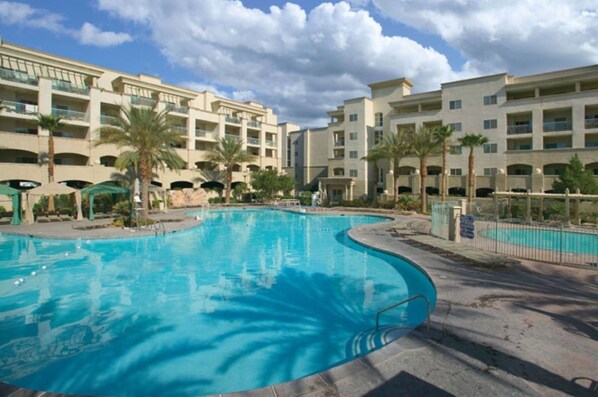 A swimming pool with a curved edge, surrounded by palm trees and a multi-story building with balconies. - Las Vegas | Group Vegas Getaway | Pool + Hot Tub | 2BR (Las Vegas,)