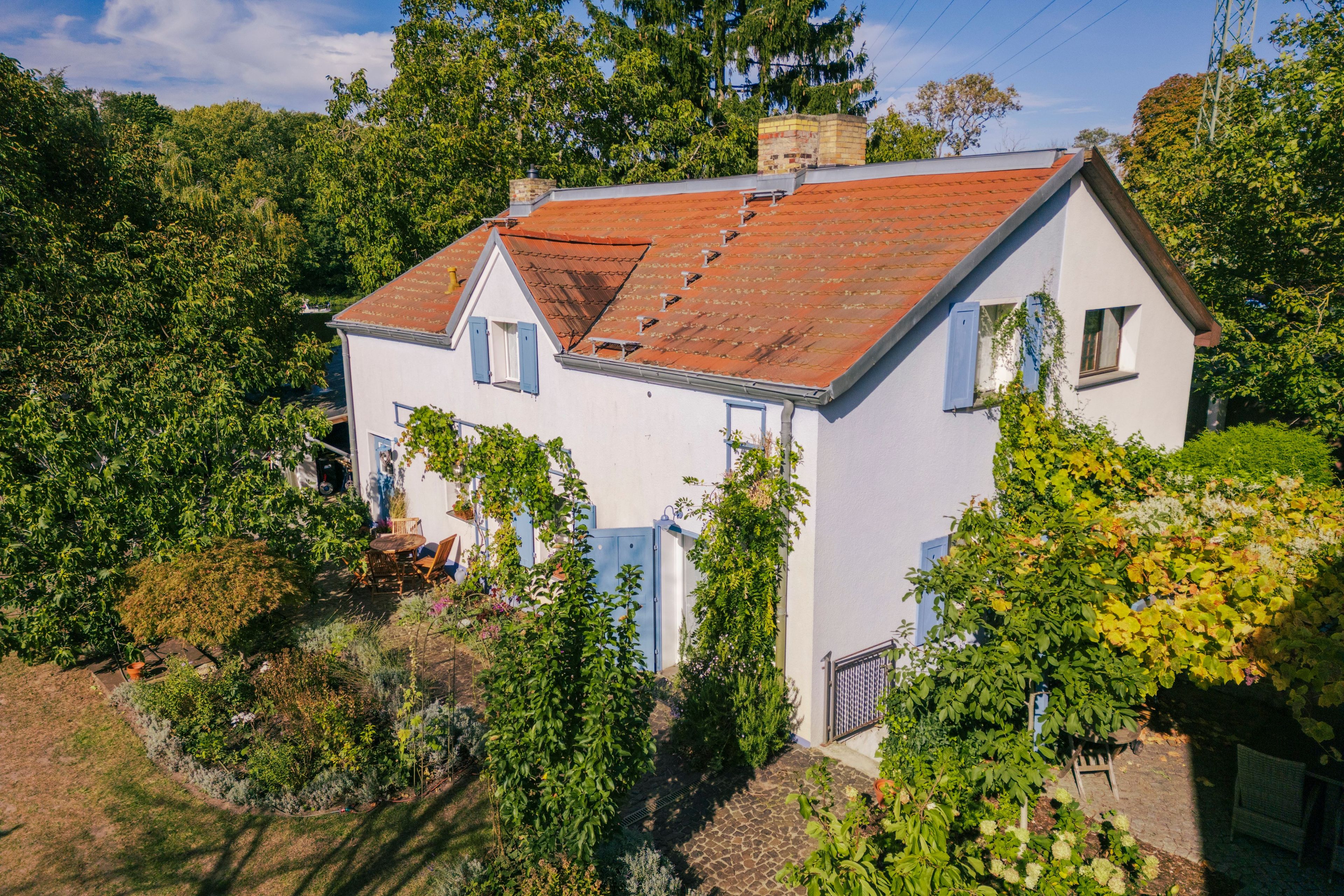 A two-story house with a red-tiled roof, surrounded by lush greenery and trees.