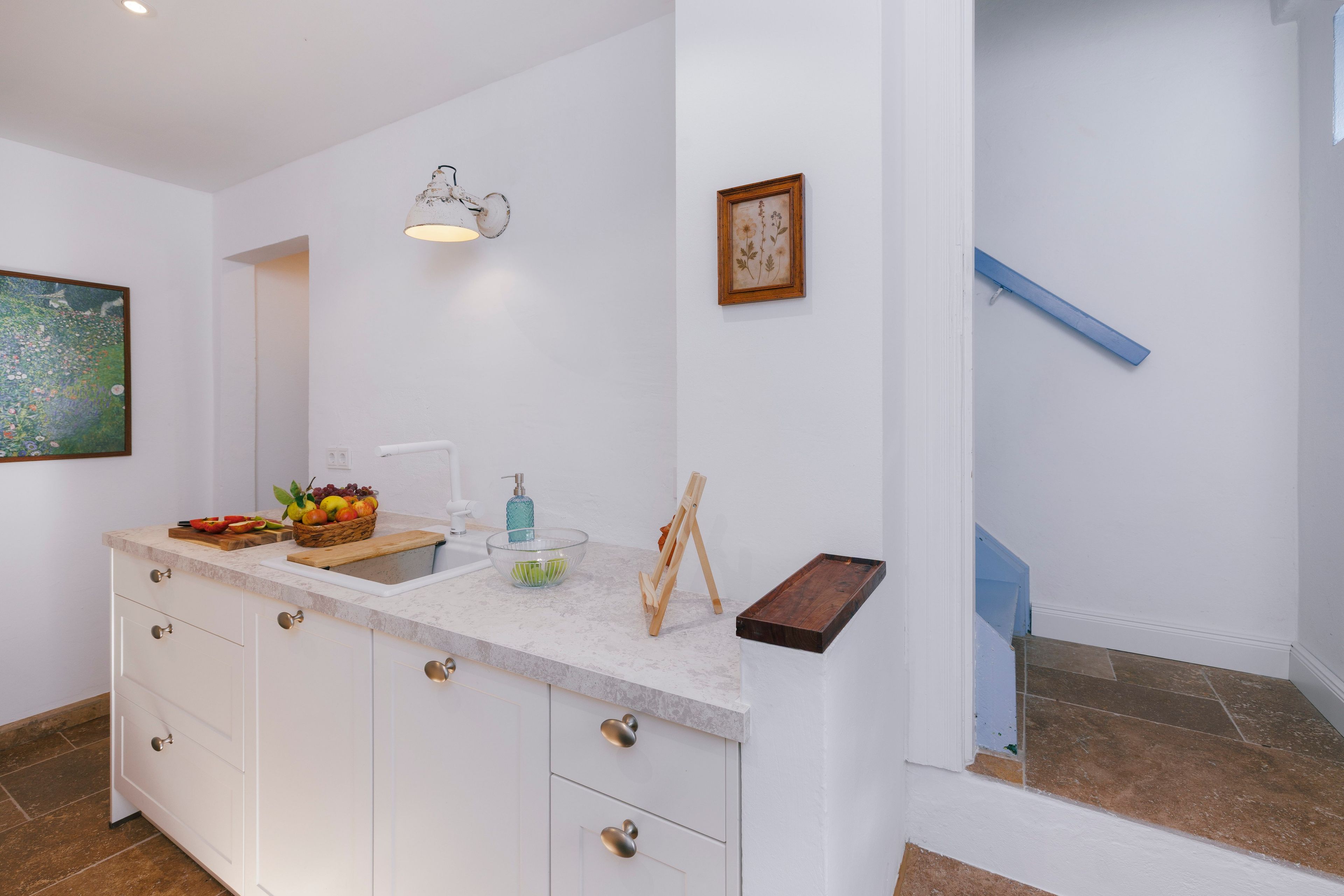 Eine moderne Küche mit Marmorarbeitsplatte, Obstkorb, Seifenspender und einem gerahmten botanischen Druck an der Wand.