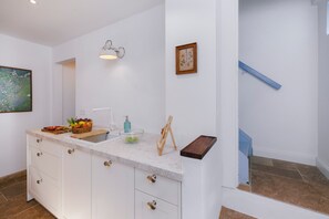 A modern kitchen with a marble countertop, a fruit basket, a soap dispenser, and a framed botanical print on the wall.