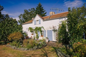 Ein zweigeschossiges Haus mit weißer Fassade, einem Ziegelkamin und einem Garten mit verschiedenen Pflanzen und einem kleinen Teich.