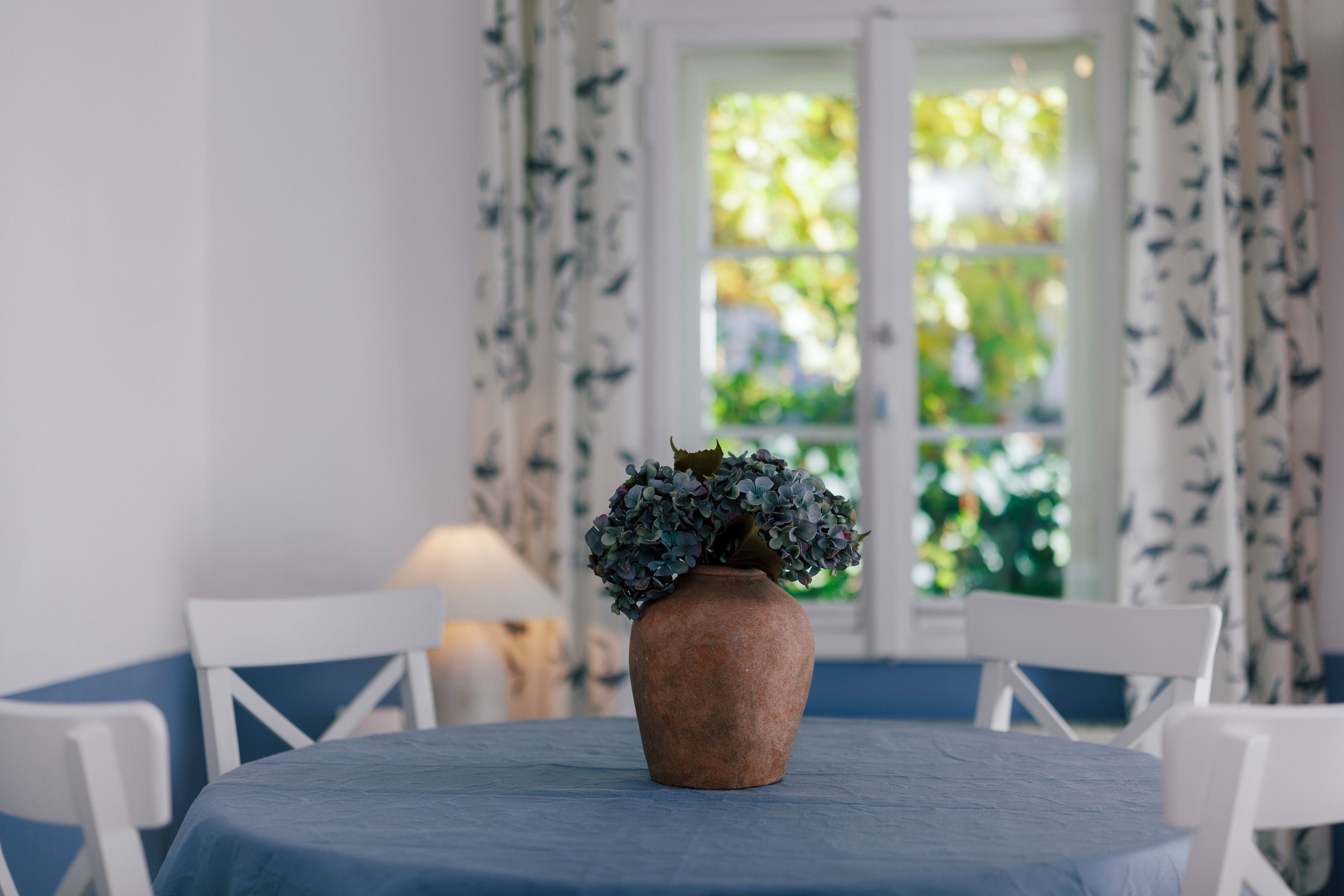A round table with a blue tablecloth, a brown vase with green foliage, and white chairs with blue cushions.