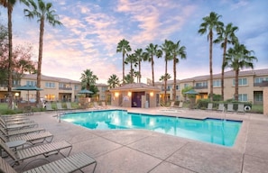 Un hotel con piscina, palmeras y una zona cubierta con sillones de descanso.