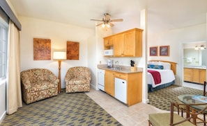 Habitación de hotel compacta con kitchenette, dos sofás estampados, zona de comedor y cama con manta roja.
