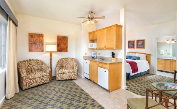 Habitación de hotel compacta con kitchenette, dos sofás estampados, zona de comedor y cama con manta roja.
