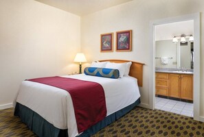 Habitación de hotel con cama, mesita de noche, lámpara, baño con lavamanos y espejo, y dos cuadros enmarcados en la pared.