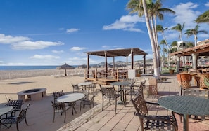 Un patio frente a la playa con mobiliario exterior, una fogata y palmeras.