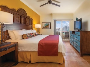 Una habitación de hotel con cama, ventilador de techo, televisión, cómoda y balcón con vistas.