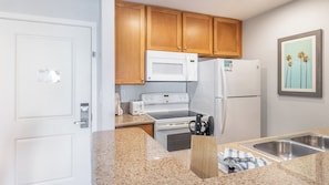 A compact kitchen with wooden cabinets, a white microwave, a white refrigerator, a stove, and a sink. - Anaheim | Romantic Disney Retreat | Pool + Parks (Anaheim)
