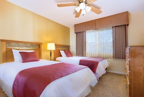 Habitación de hotel con dos camas, ventilador de techo, mesita de noche con lámpara, cómoda y ventana con cortinas.