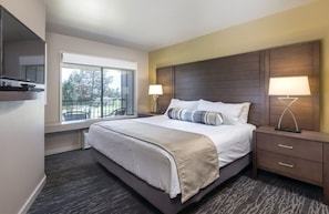 A hotel room with a large bed, two bedside tables with lamps, a window with a view of trees, and a dark wood headboard. - Redmond | High Desert Comfort Escape | Pool + Trails (Redmond)
