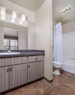 A bathroom with a wooden vanity, a white toilet, a bathtub, and a shower curtain. - Redmond | High Desert Comfort Escape | Pool + Trails (Redmond)
