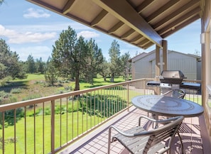 A balcony with a grill, a table, and chairs, overlooking a grassy area with trees. - Redmond | High Desert Easy Access Stay | Pool + Trails | 2BR Ground Floor (Redmond)