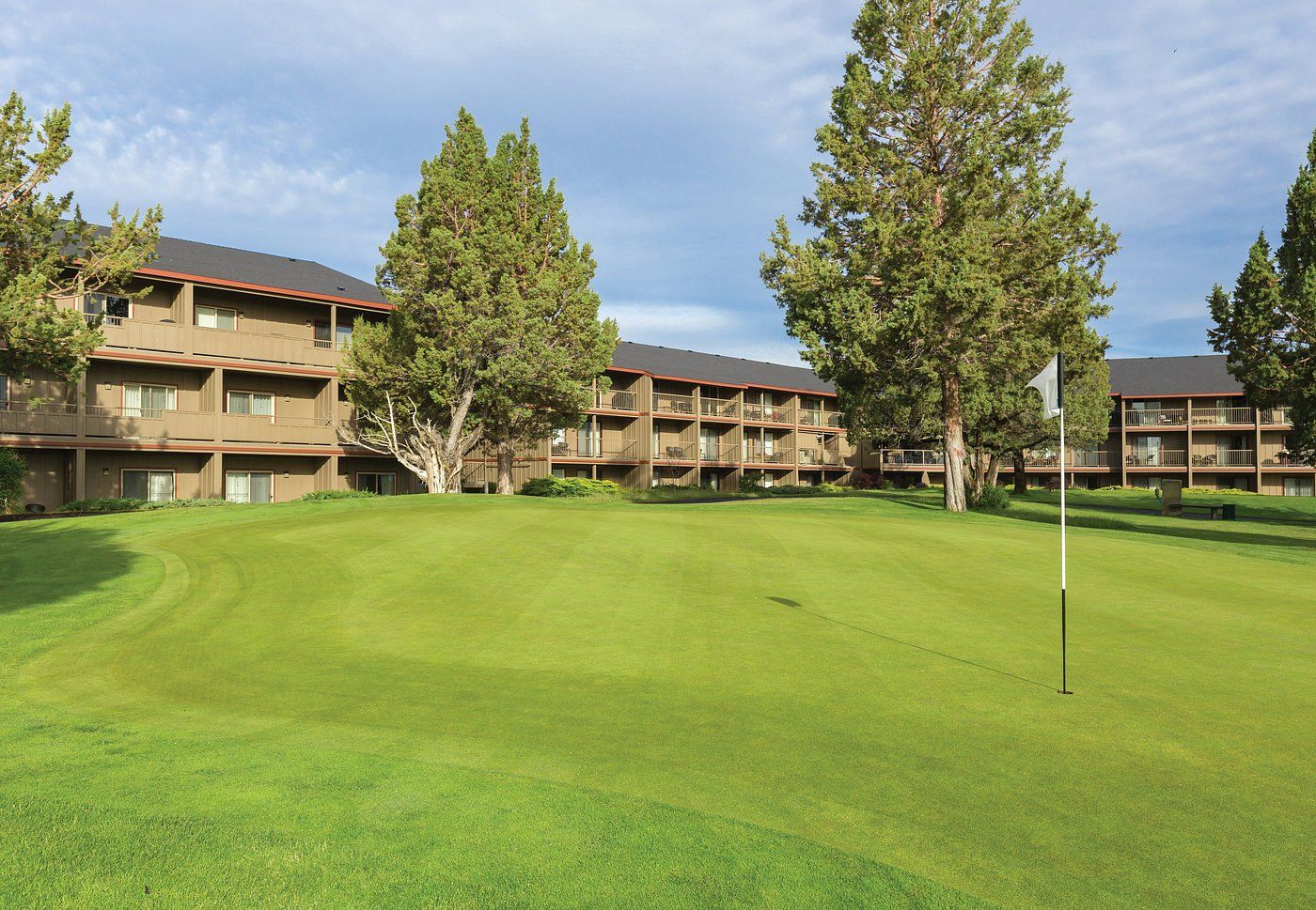 A golf course with a putting green, surrounded by multi-story buildings with balconies and large trees.