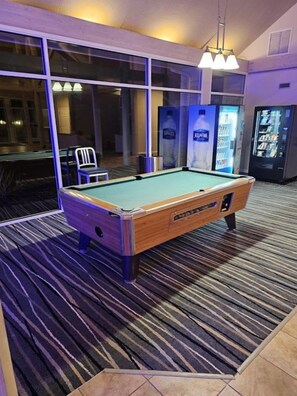 Una mesa de billar en una sala con ventanas amplias, una silla y máquinas expendedoras.