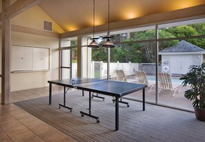 Mesa de ping pong en una habitación con ventanas amplias, una pizarra y una piscina visible a través de las ventanas.