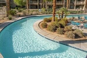 A curved pool with clear water, surrounded by a landscaped area with small bushes and palm trees. There are lounge chairs and a fence visible in the background. - Indio | Desert Relaxation Stay | Pool & Lazy River (Indio)