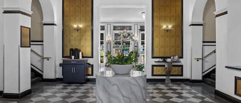 A lobby with a marble column, checkered floor, chandeliers, and a view into a room with a large window.