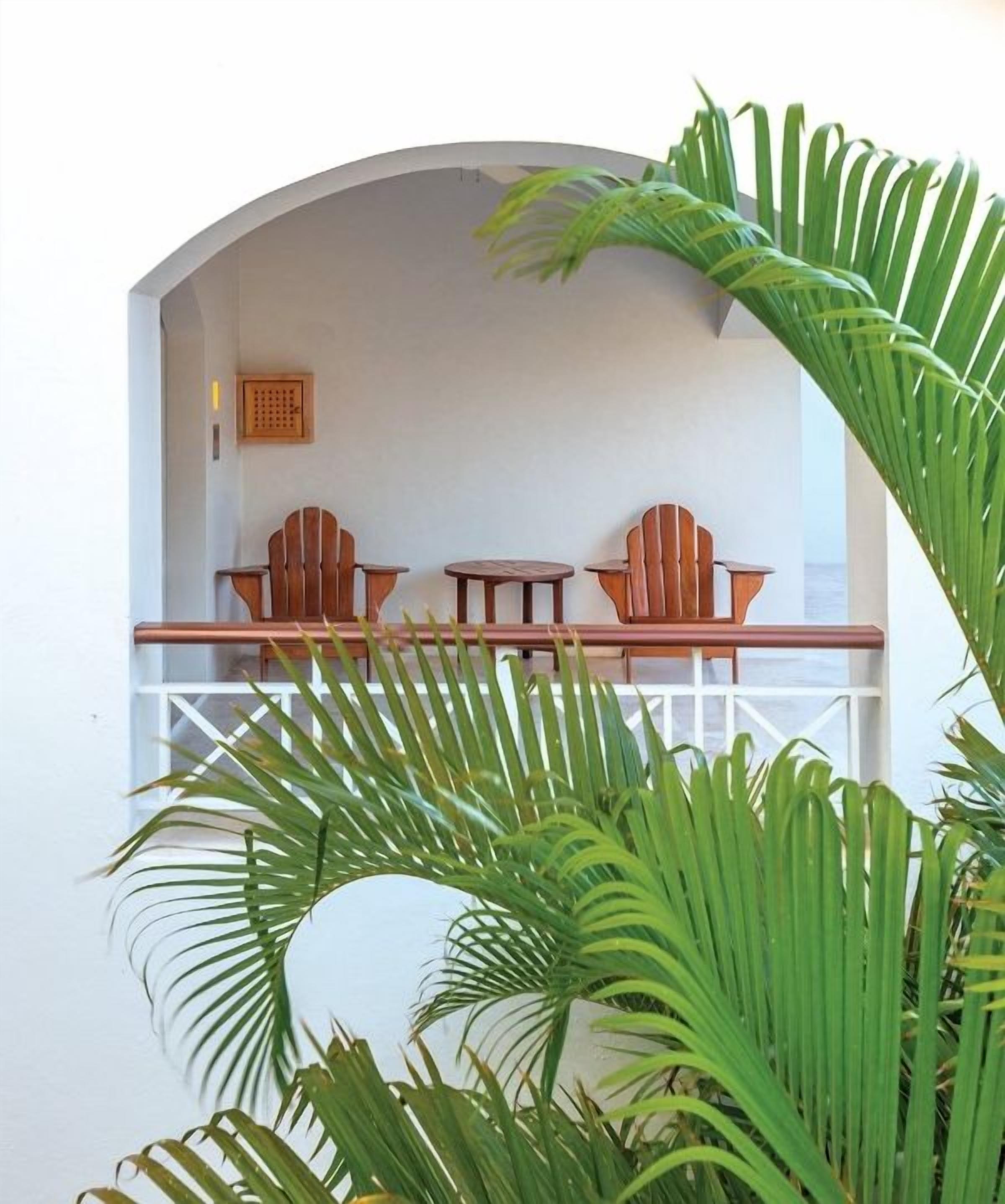 An arched balcony with wooden chairs and a table, surrounded by lush green foliage.