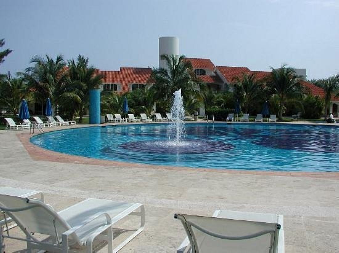 A swimming pool with a fountain, surrounded by lounge chairs and palm trees.