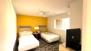 Habitación de hotel con dos camas, ventilador de techo, televisión y ventana con persianas.