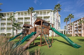 Un parque infantil con toboganes y estructuras de escalada frente a un hotel con múltiples balcones y palmeras.