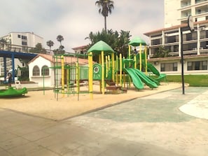 A playground with climbing structures and slides, surrounded by buildings and palm trees. - Rosarito | Family Beach Retreat | Pool + Beach | 2BR (Rosarito)