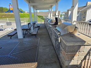 Eine Terrasse im Freien mit Grillanlagen, Picknicktischen und einem Spielplatz im Hintergrund.