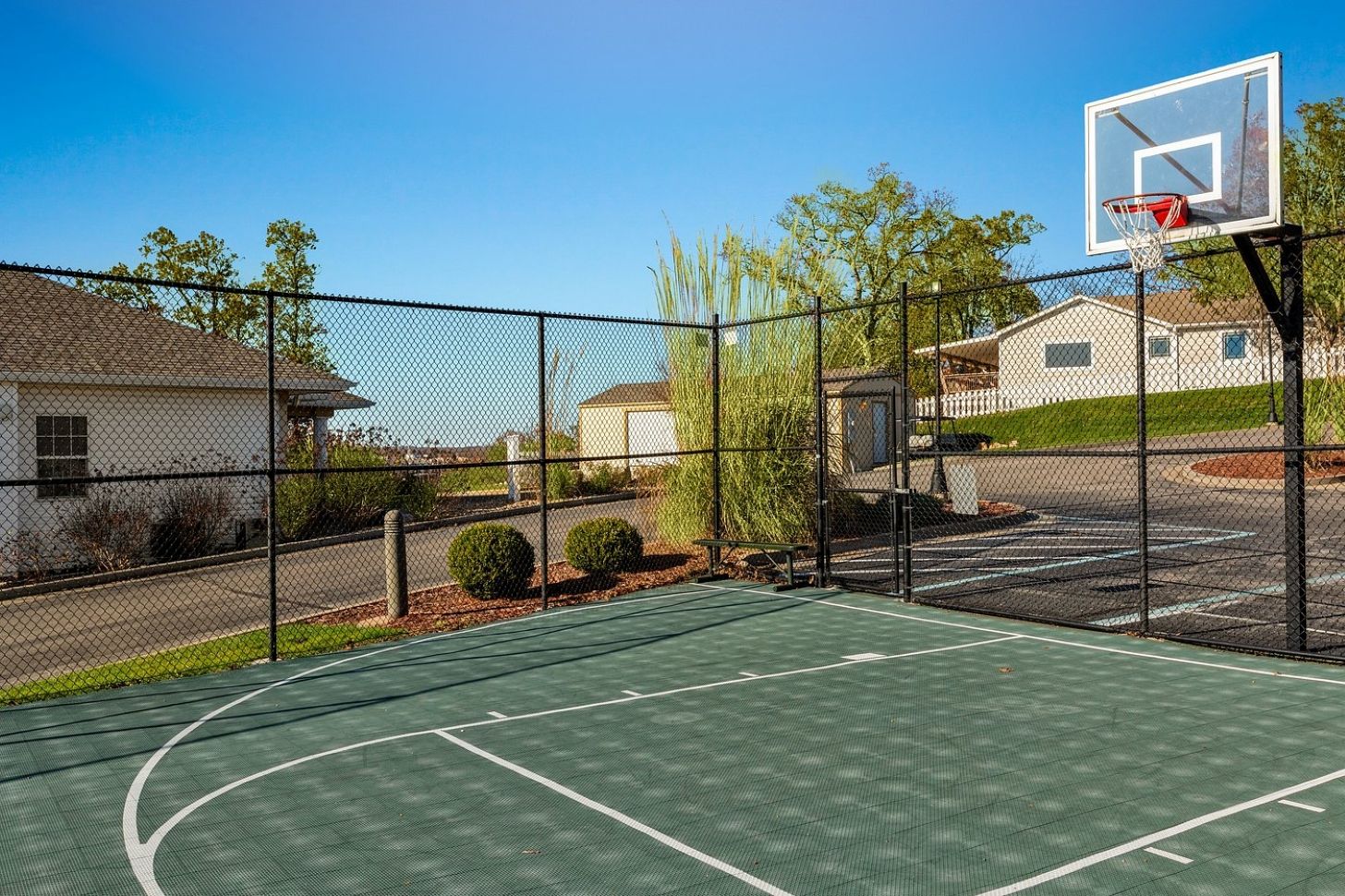 Una cancha de baloncesto al aire libre con una valla de malla, un aro de baloncesto y casas al fondo.