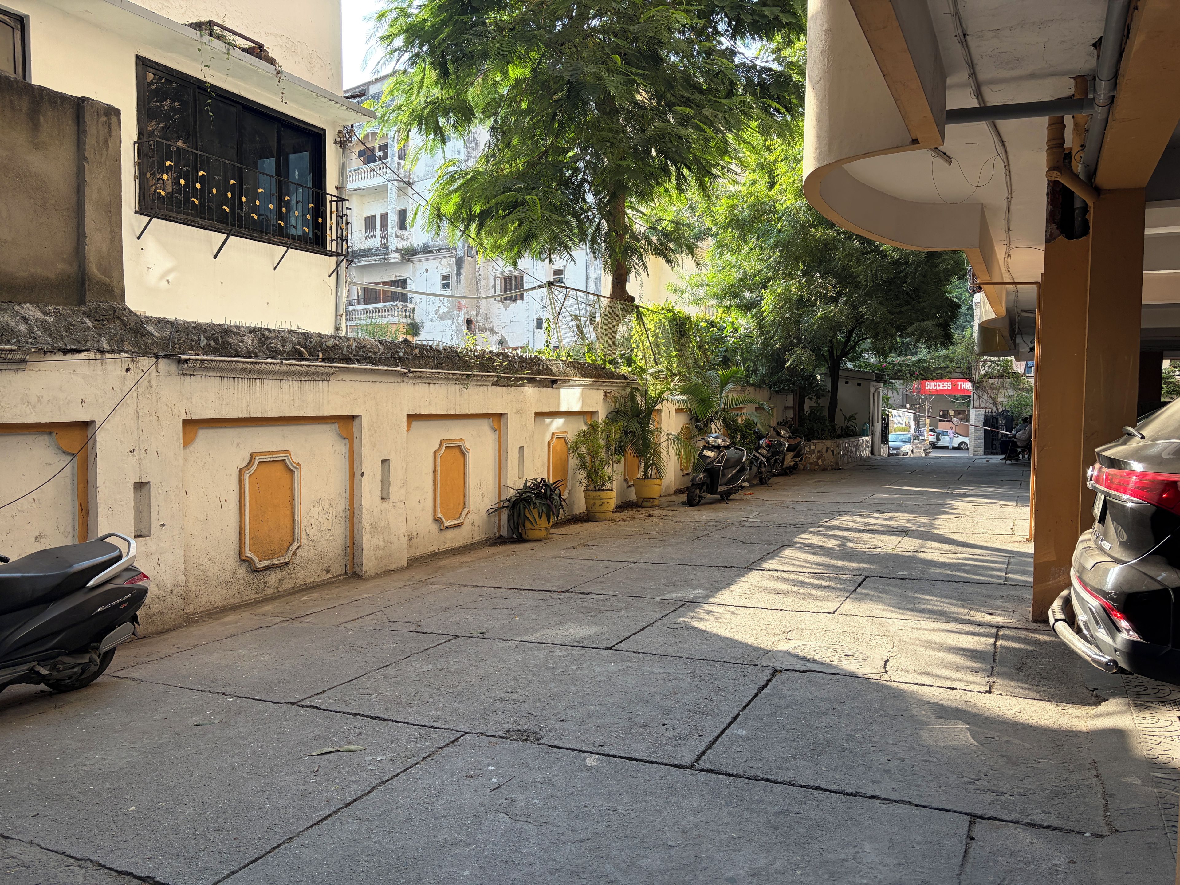 Una calle estrecha con scooters y un coche aparcados, edificios con balcones y plantas en macetas a lo largo de la acera.