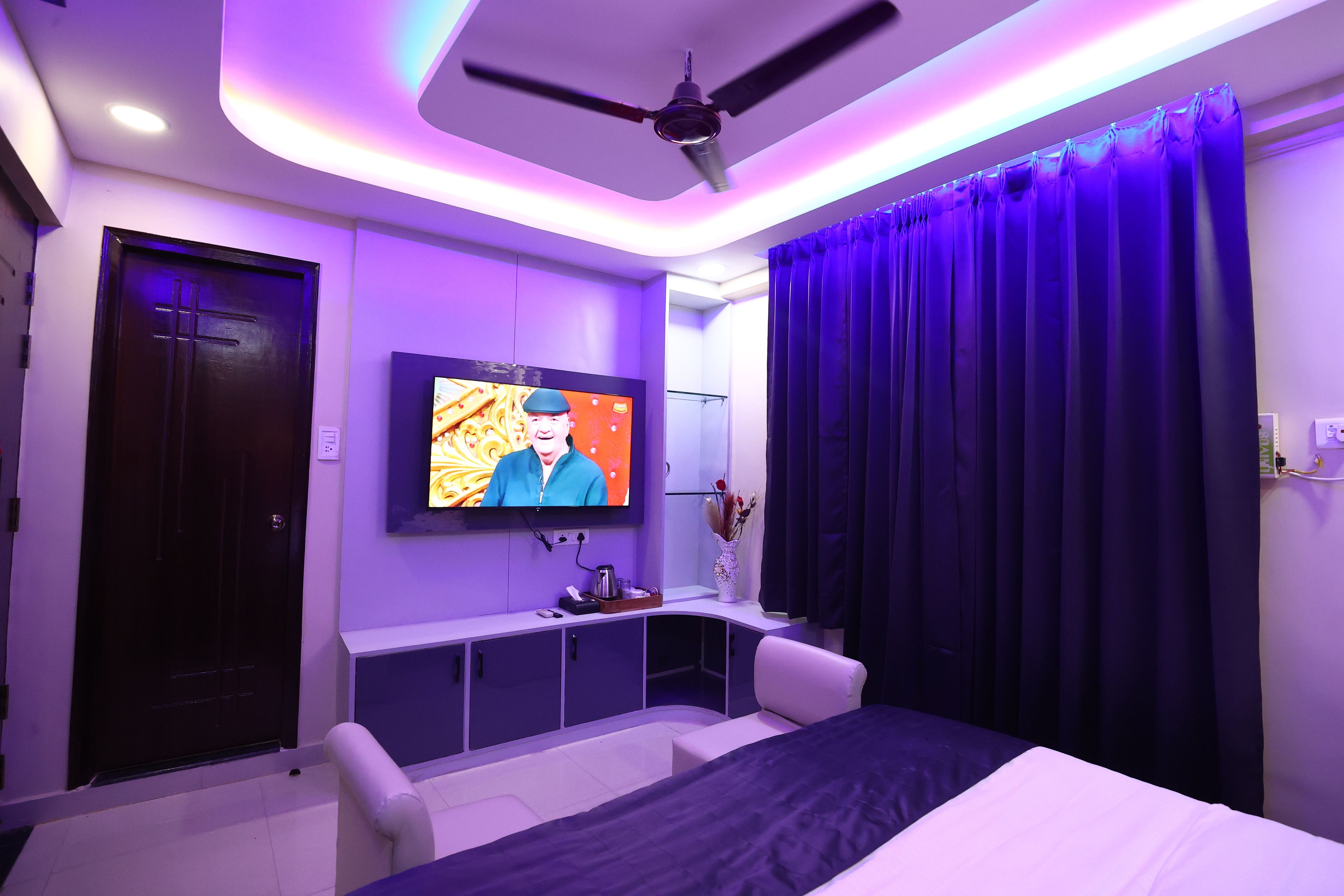 Un dormitorio moderno con una cama, un televisor montado en la pared, un ventilador de techo y una puerta de color oscuro.