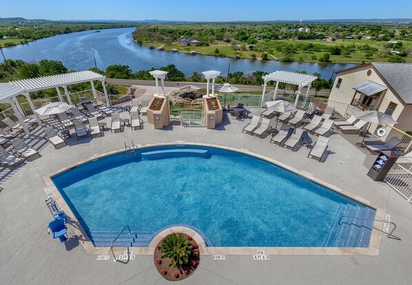 A swimming pool with lounge chairs and umbrellas, surrounded by a landscaped area with a view of a river and hills. - Marble Falls | Premium Lake Retreat | Pool + Lake | 2BR (Marble Falls)