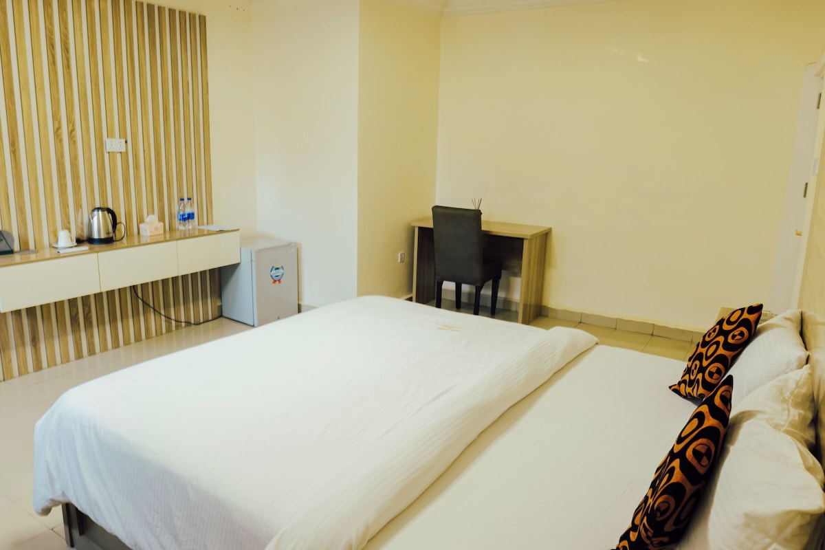 A hotel room with a bed, a desk, a chair, a minibar, and a wall-mounted shelf with a kettle and bottles.