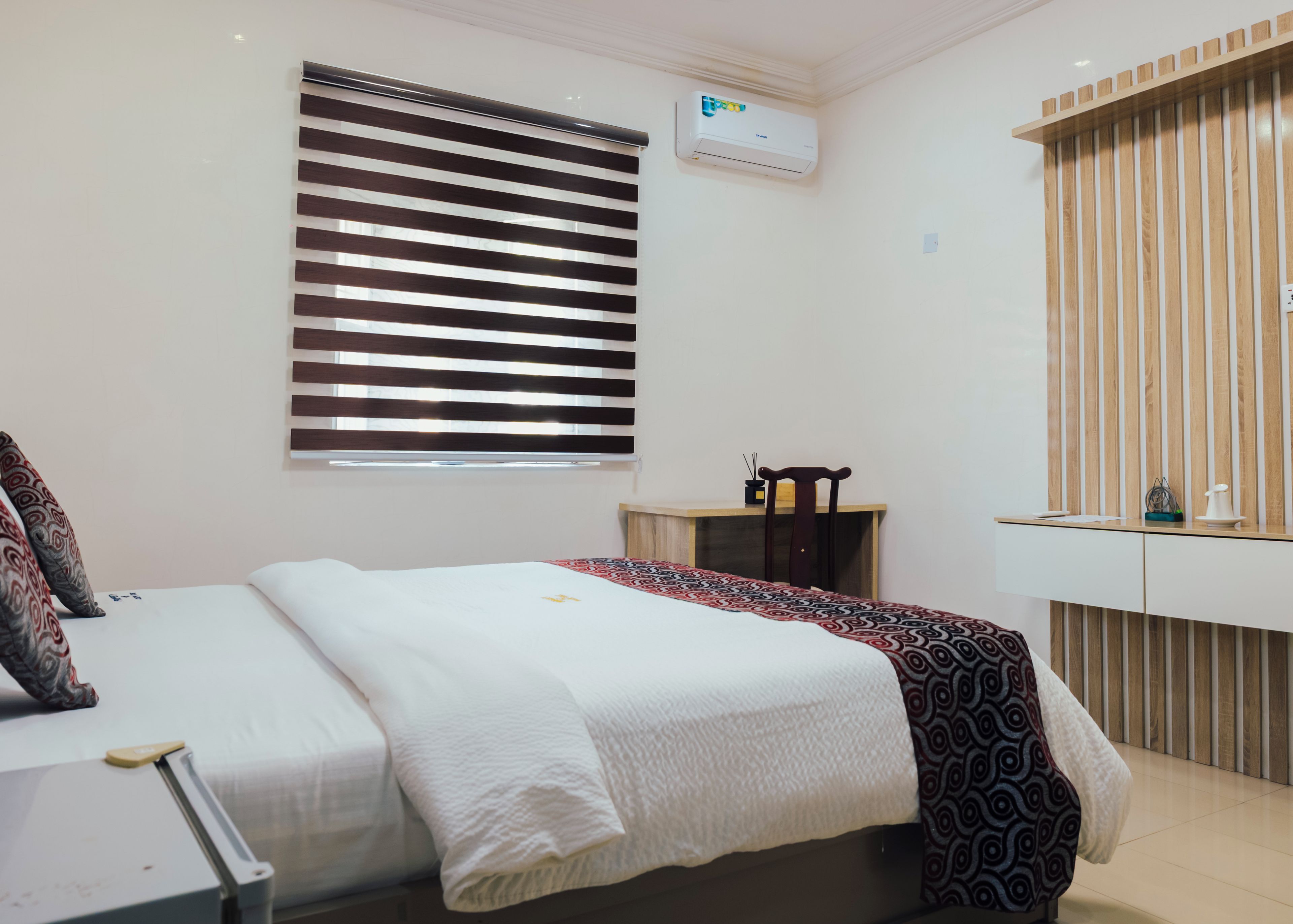 Un dormitorio con cama, escritorio, silla, ventana con persianas y aire acondicionado.