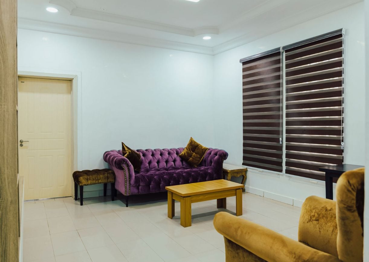 Ein Zimmer mit einem violetten Sofa, einem Holztisch und einem Fenster mit Jalousien.