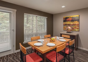 Une salle à manger avec une table mise pour six personnes, des chaises avec des coussins rouges, un panier de fruits et un tableau accroché au mur.