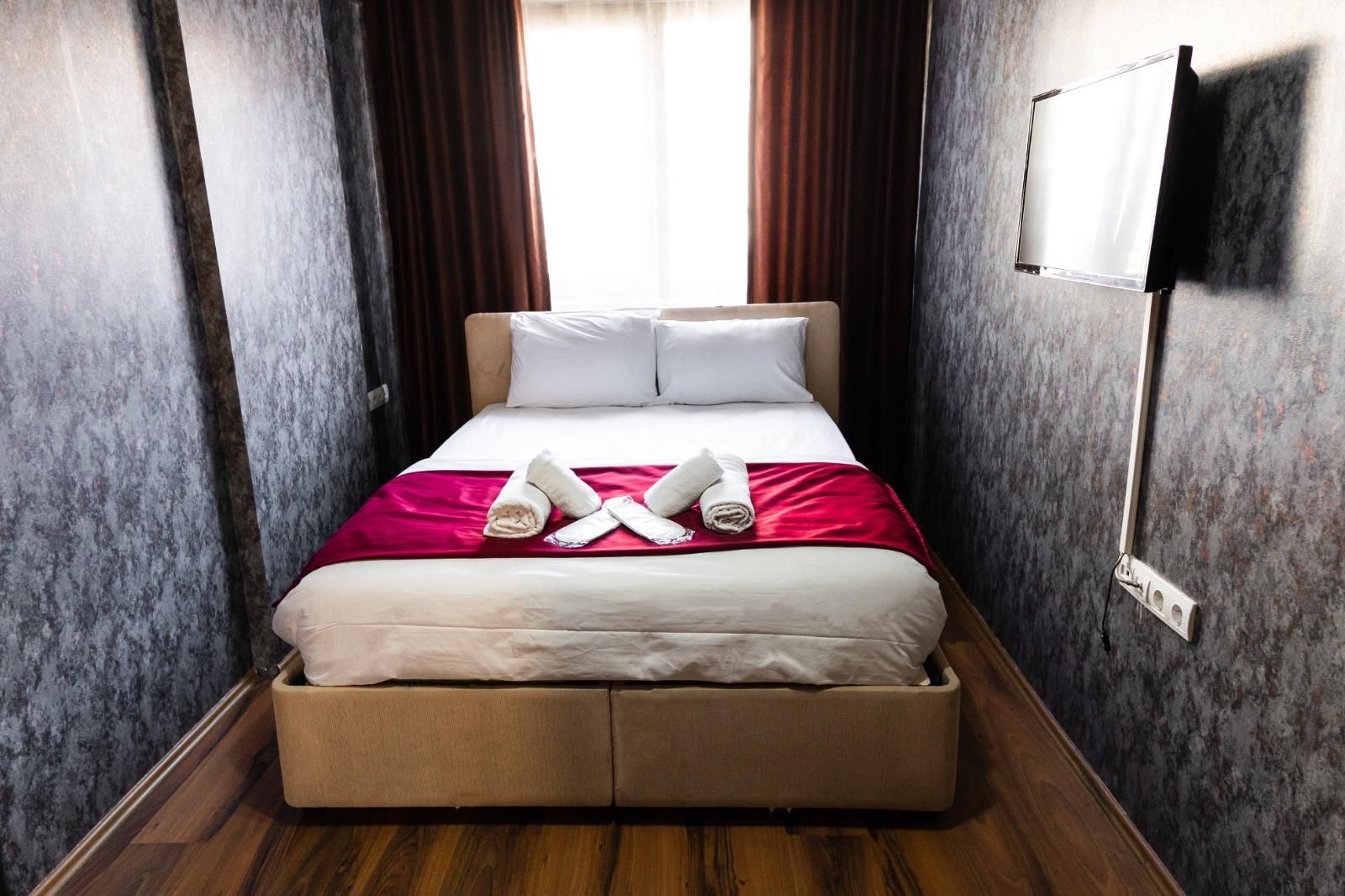 Une chambre d’hôtel avec un lit, un plaid rouge, deux serviettes enroulées et un téléviseur fixé au mur.