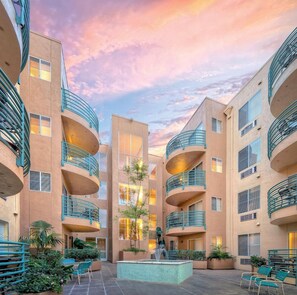 Un immeuble résidentiel à plusieurs étages doté de balcons, d’une cour intérieure avec des sièges et une fontaine, et d’un ciel au coucher du soleil.