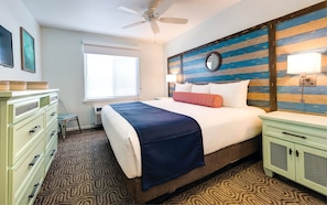 Une chambre d’hôtel comprenant un lit, une table de chevet, une commode, un ventilateur de plafond, une fenêtre avec des stores et un mur à motifs rayés.