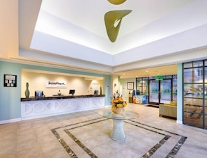 Ein Hotel-Empfangsbereich mit Empfangstresen, einem Glastisch mit Blumenvase und einer Sitzecke mit Sofa und Sesseln.