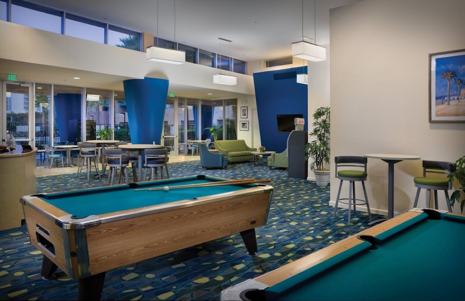 A pool table in a hotel lobby with a view of the outdoors through large windows.