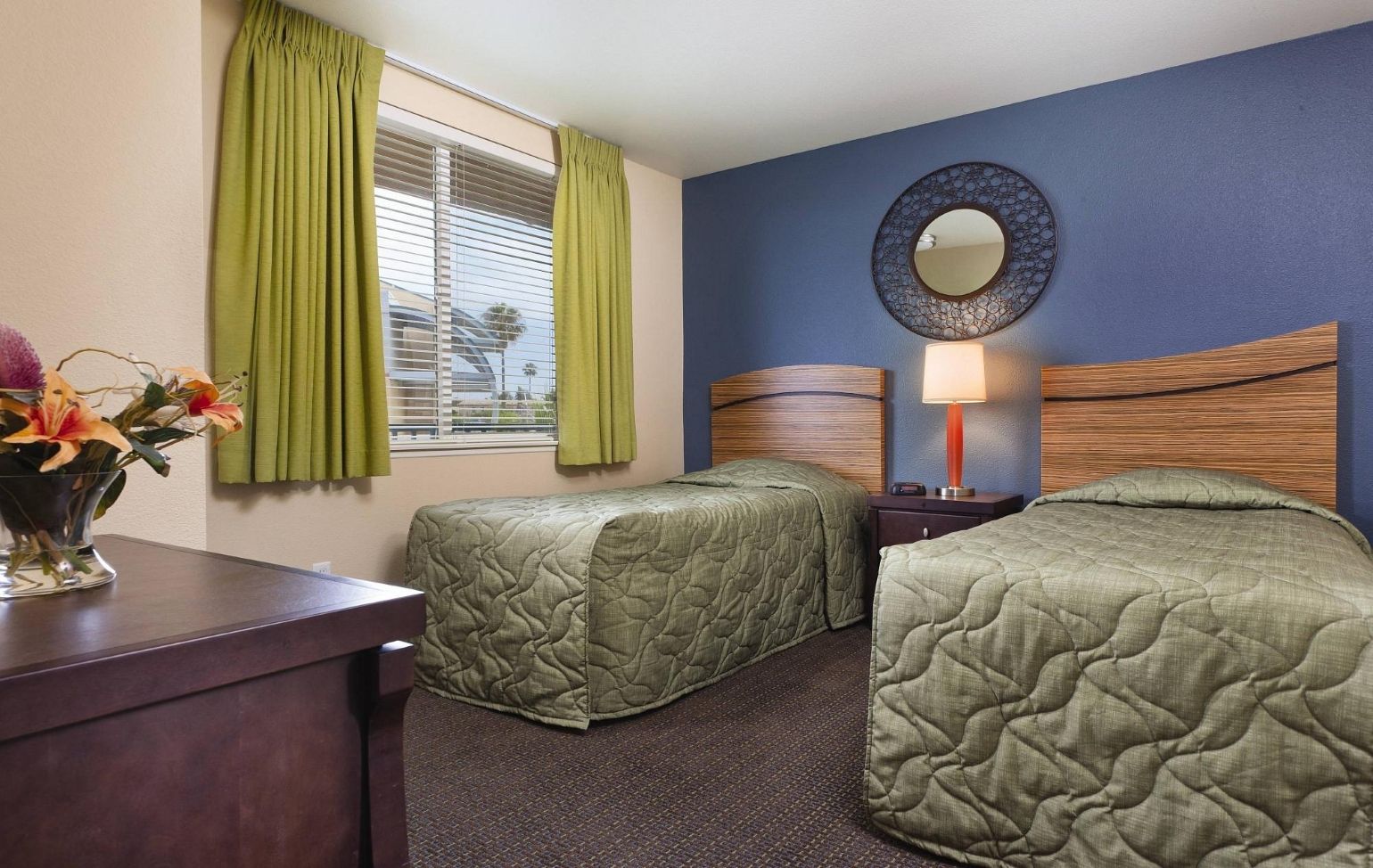 A hotel room with two beds, a nightstand with a lamp, a mirror on the wall, and a window with curtains.