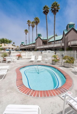 A swimming pool with a brick border, surrounded by white lounge chairs, palm trees, and a white picket fence.