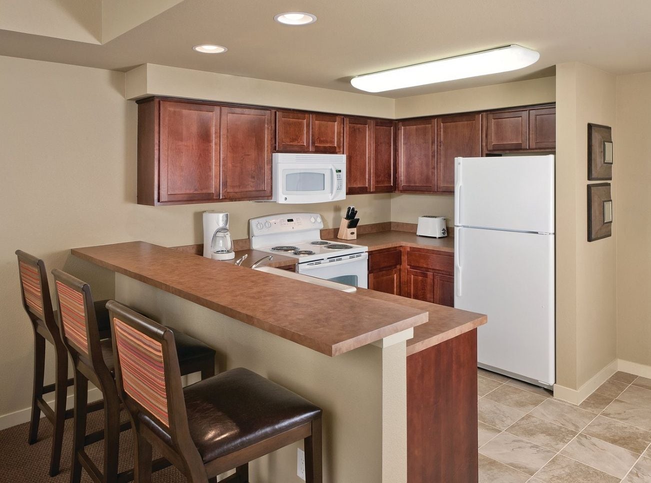 A kitchen with wooden cabinets, a white refrigerator, a stove, and a microwave. There are three bar stools and a coffee maker.