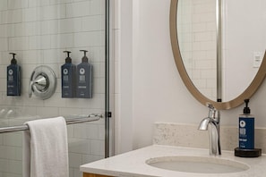 Une salle de bain avec une douche en verre, un lavabo doté d’un robinet moderne, un miroir avec un cadre doré et des articles de toilette fixés au mur.